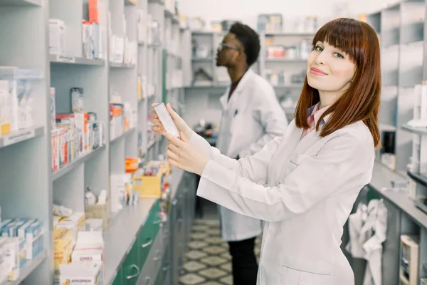 Two Pharmacists Working. Caucasian Woman and African Man Wearing Special Medical Uniform. Woman Showing package with Pills. Man looking for medicines on Background.