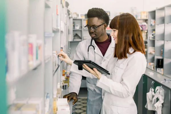 Confident Male And Female Pharmacists In Pharmacy. female pharmacist talking with her colleague about the attributes and side effects of a medicine or pharmaceutical product for sale, in the pharmacy