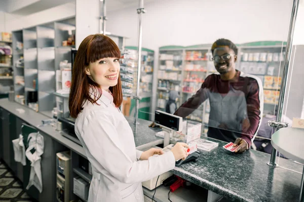 Felnőtt afrikai férfi ügyfél kérdezi a fiatal kaukázusi nő gyógyszerész a gyógyszerek közelében Cashbox a patikus — Stock Fotó