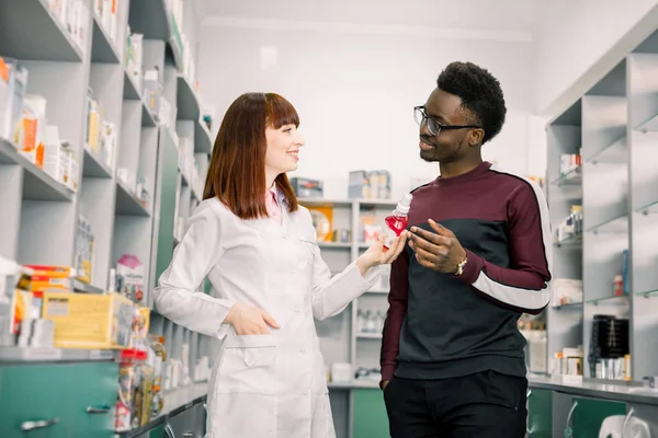Krásná ženská lékárnice mluví se samečkem Afričanem a přitom mu ukazuje láhev Ústní sprej. — Stock fotografie