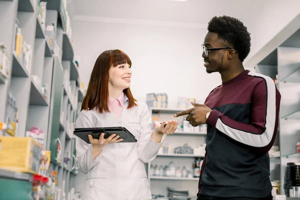 Medicine, healthcare and people concept - young woman apothecary with drug and African male customer at pharmacy — Stock Photo, Image