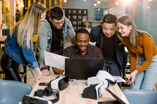 Groupe de collaborateurs adultes de Hipsters utilisant des gadgets modernes. Quatre amis d'affaires, souriants et gestuels pendant que leur ami africain travaille sur un ordinateur portable à la table . — Photo