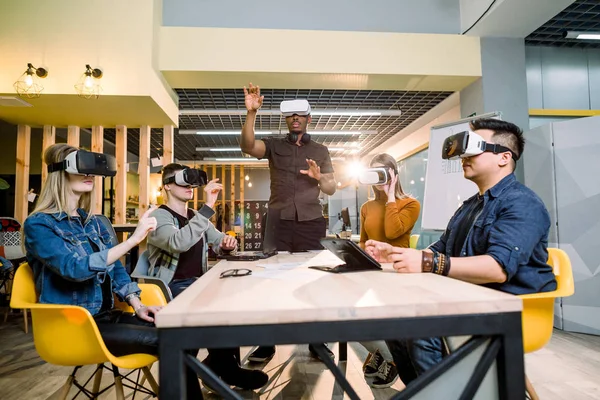 Enjoying new experience. Handsome young African man in VR headset gesturing and smiling while standing at the table. Young people wearing vr goggles