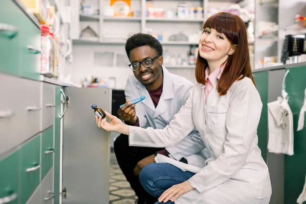 Boční pohled na dva specializované lékárníky, Afričana a kavkazskou ženu, hledaje nejlepší medicínu při práci v komunitní lékárně — Stock fotografie