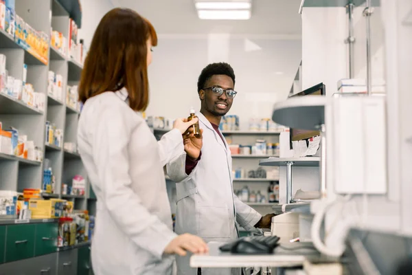 medicine, pharmaceutics, health care and people concept - happy African man pharmacist showing bottle of drug to young Caucasian woman pharmacist at drugstore