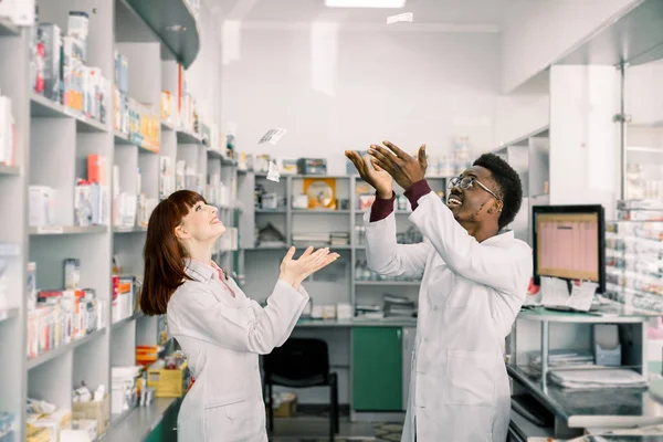 Dva legrační usmívající se mladí lékárníci, kteří se baví v lékárně, zahazovat různé prášky — Stock fotografie