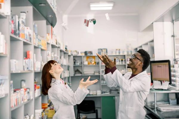 Dva veselí multiethičtí lékárníci, muži a ženy spolupracují a baví se, házeli prášky na puchýři. Moderní lékárna — Stock fotografie