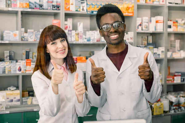 약국에 오신 것을 환영합니다. 행복한 미소 두 약사, 아프리카 남자와 백인 여자, 엄지 손가락을 제스처, 현대 약국에 서있는 동안 — 스톡 사진