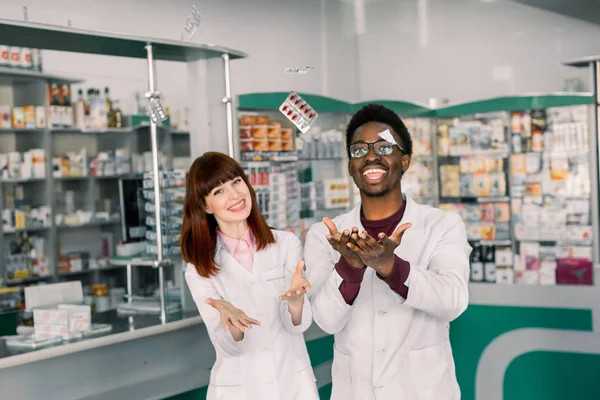 Két vidám multietnikus gyógyszerészek, férfi és nő dolgozik együtt, és jól érzik magukat, hányt hólyagok tablettát. Modern gyógyszertár — Stock Fotó