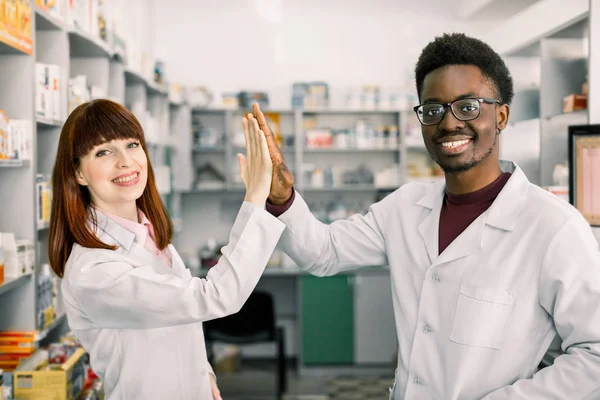 Dois farmacêuticos de coleções felizes, homem africano e mulher caucasiana que trabalha na farmácia, sorrindo. dando cinco e se divertir . — Fotografia de Stock