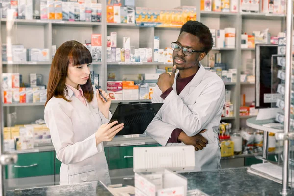 Retrato de farmacêutico Africano masculino em pé com colega do sexo feminino com área de transferência, moderno fundo farmácia — Fotografia de Stock