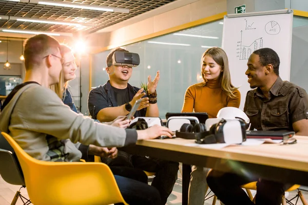 Travail d'équipe essayant des lunettes de réalité virtuelle pour travailler dans un bureau créatif. Jeune homme asiatique en utilisant de nouvelles technologies vr lunettes tandis que ses collègues parlent assis à la table — Photo