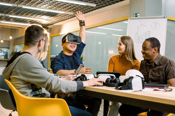 Fem unga roliga människor sitter vid bordet framför varandra, en man använder Virtual Reality Goggles. VR Goggles koncept, affärsmöte — Stockfoto