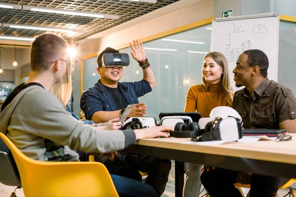 Five young funny people sitting at the table in front of each other, one man is using virtual reality goggles. VR goggles concept, business meeting
