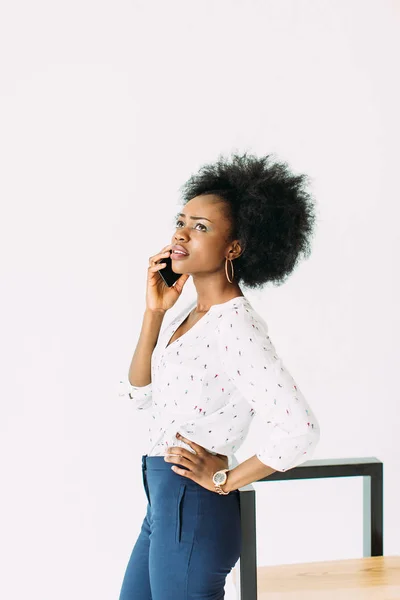 Allegra giovane donna d'affari afro-americana che parla al cellulare, in piedi vicino alla sedia moderna, isolata sul bianco — Foto Stock