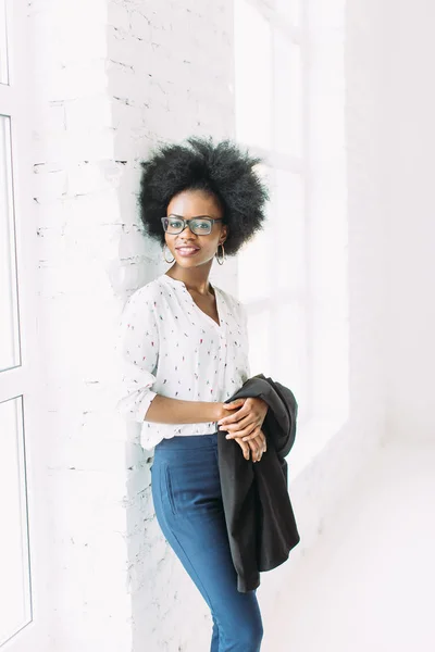 Young african american business woman in glasses, standing near the big window and holding jacket