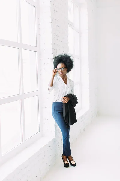 Jonge African American Business vrouw in glazen, staande in de buurt van het grote raam en het houden van jas — Stockfoto