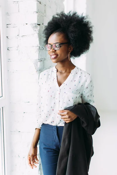 Glimlachend jonge Afro Amerikaanse zakenvrouw met een jas, staande in de buurt van het grote raam in Studio — Stockfoto