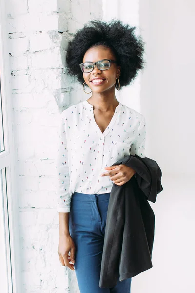 Glimlachend jonge Afro Amerikaanse zakenvrouw met een jas, staande in de buurt van het grote raam in Studio — Stockfoto