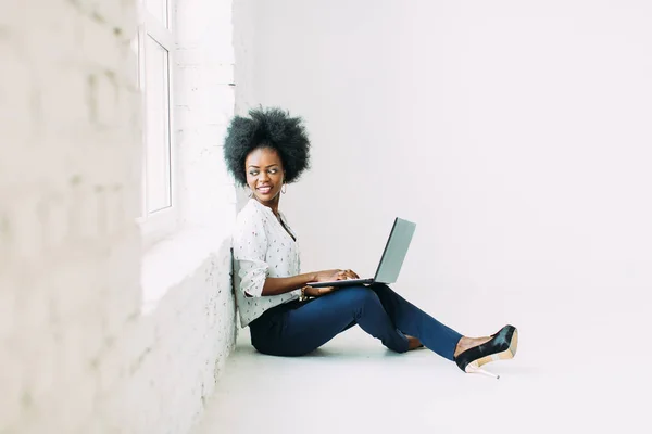 Giovane donna d'affari afroamericana che utilizza il computer portatile, mentre seduto sul pavimento vicino a una grande finestra in studio — Foto Stock