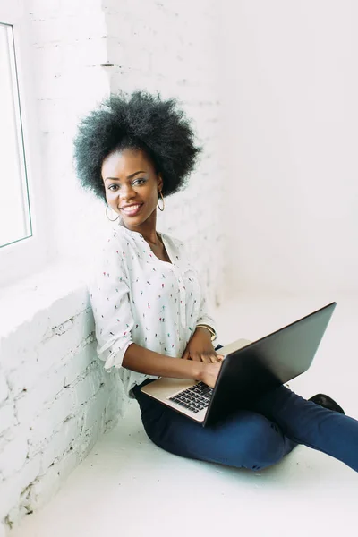 Giovane donna d'affari afro-americana sorridente che utilizza il computer portatile, seduta sul pavimento in uno studio di luce bianca — Foto Stock