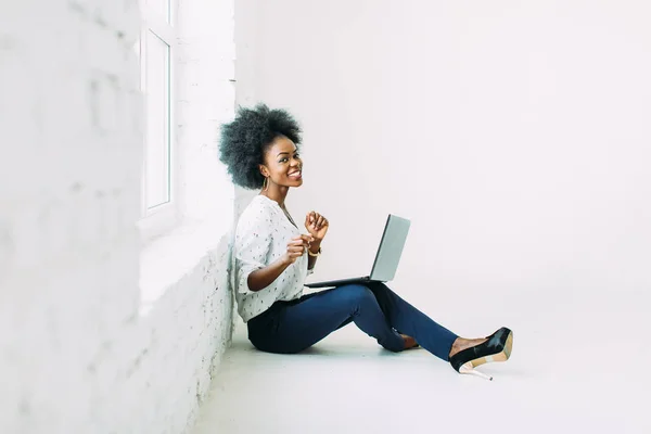 Giovane donna d'affari afroamericana che utilizza il computer portatile, mentre seduto sul pavimento vicino a una grande finestra in studio — Foto Stock