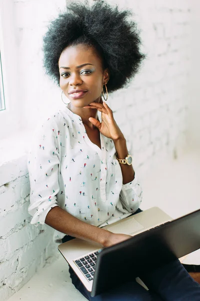 Giovane donna d'affari afro-americana sorridente che utilizza il computer portatile, seduta sul pavimento in uno studio di luce bianca — Foto Stock