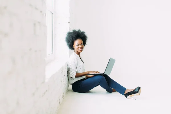 Giovane donna d'affari afroamericana che utilizza il computer portatile, mentre seduto sul pavimento vicino a una grande finestra in studio — Foto Stock