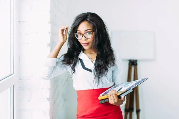 Mulher de negócios afro-americana ou professora de óculos segurando livros e documentos, de pé sobre fundo branco perto de uma grande janela — Fotografia de Stock