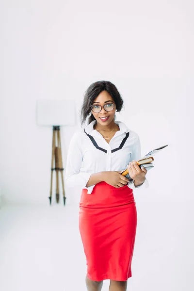 Bella giovane africana sorridente insegnante donna in camicia bianca e gonna rossa in possesso di libri e documenti — Foto Stock