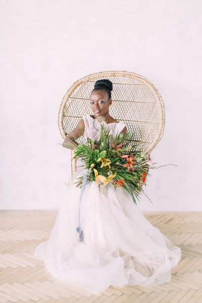 Bela noiva de pele escura. Linda noiva afro-americana com buquê tropical em estúdio, sentada na cadeira — Fotografia de Stock