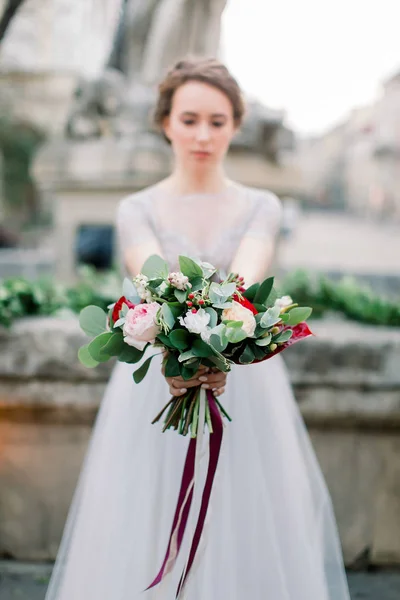 Retrato de novia hermosa en vestido de novia increíble celebración de ramo tierno, mientras que de pie al aire libre en el fondo del monumento de piedra en el centro de la ciudad vieja. Concéntrate en el ramo —  Fotos de Stock