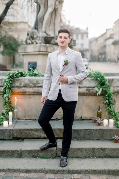 Stylowy uśmiechnięty Pan młody w szarym płaszczu, pozowanie na zewnątrz, stojąc na kamiennych schodach pomnika w centrum starego miasta przed ceremonią ślubną — Zdjęcie stockowe
