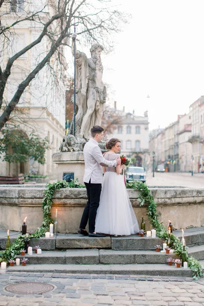 Romantico matrimonio coppia nella parte antica della città Lviv. Elegante sposo che abbraccia la sua sposa dal retro, in piedi sulle scale di pietra del monumento . — Foto Stock