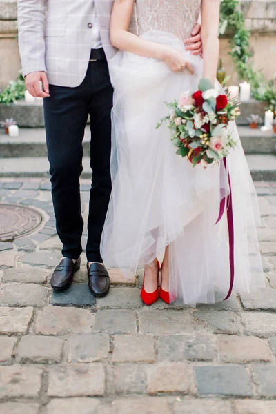 Nohy stylové svatební dvojice, ženich v černých botách a nevěstě v červených botách s luxusními svatební šaty, držící svatební kytici. Venkovní záběr ve starověkém městě. — Stock fotografie