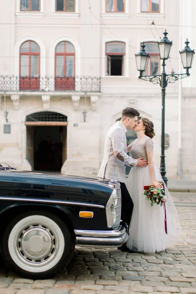 Felice matrimonio di lusso coppia baciare e abbracciare vicino nero auto retrò nel centro storico della città, antichi edifici sullo sfondo . — Foto Stock