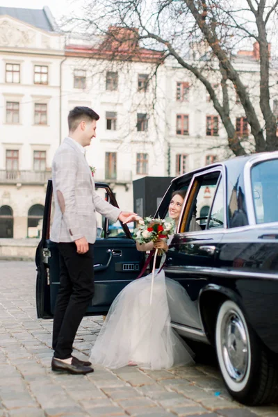 Krásná žena na svatebních šatech, která sedí ve stylovém černém retro autě a pohledný ženich, který dává ruku své nevěstě. Staré městské budovy na pozadí — Stock fotografie