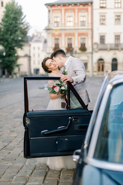 Novia con novio cerca del coche viejo. Recién casados besándose y abrazándose mientras están de pie detrás de un viejo coche retro negro en el centro de la ciudad vieja. Lviv, Ucrania — Foto de Stock