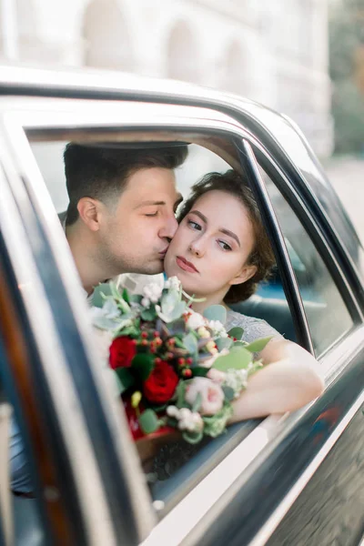 Sposa con sposo seduto in vecchia auto retrò nera. Sposi baciare e abbracciare mentre seduto all'interno della vecchia auto retrò nero nel centro storico della città. Lviv, Ucraina — Foto Stock