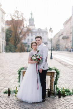 Muhteşem gelin ve yakışıklı damat ayakta ve arka planda çiçek ve mumlar ile vintage dekore ahşap masa yakınında sarılma