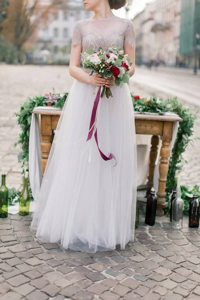 Hermosa novia sostiene un ramo de novia, de pie cerca de la mesa de madera con una decoración en el centro del casco antiguo —  Fotos de Stock