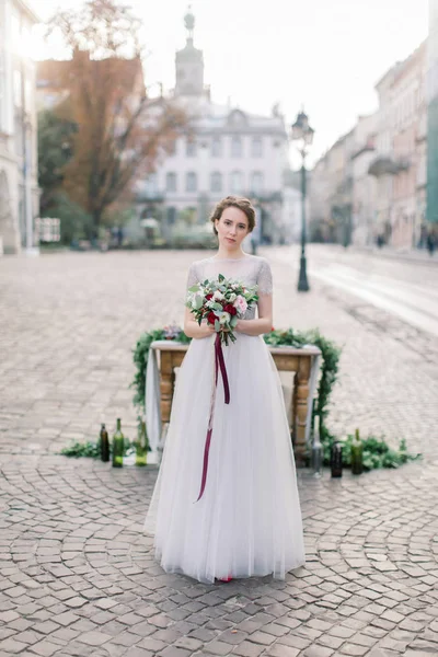 Krásná nevěsta má svatební kytici stojící nedaleko dřevěného stolu s výzdobou uprostřed starého města — Stock fotografie