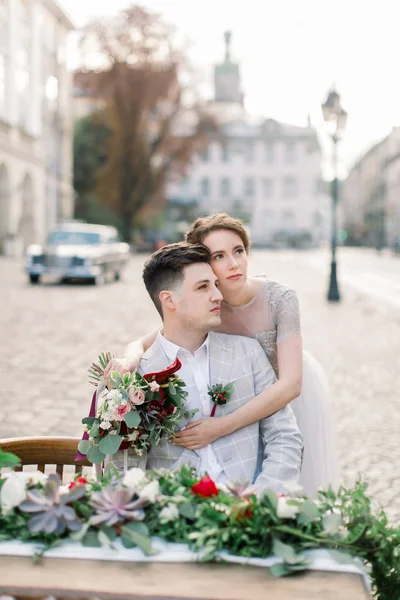 Güzel gelin ve yakışıklı damat yakın çekim portre, sarılma ve dekore edilmiş masada oturan, eski şehir arka plan