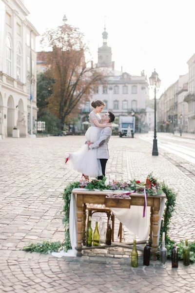 Ženich drží svou nevěstu v náručí a stojí na pozadí starých budov ve městě. — Stock fotografie