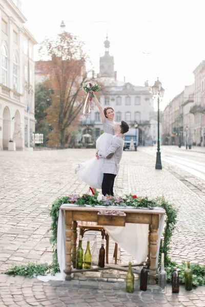Ženich drží svou nevěstu v náručí a stojí na pozadí starých budov ve městě. — Stock fotografie