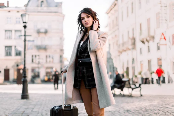 Bella giovane donna con i capelli corti scuri in abiti casual alla moda che viaggiano con una valigia a ruote, passeggiando per la strada della vecchia città europea — Foto Stock