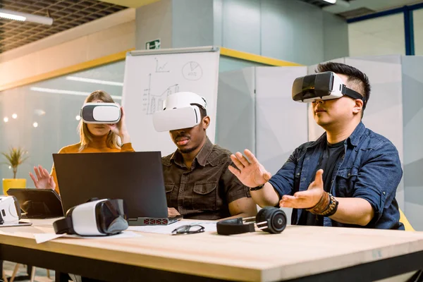Fiatal csoport multietnikus emberek szórakozik az új technológia VR headset. Kaukázusi lány, afrikai és kínai férfi ül az íróasztalon, és megpróbálta VR szemüvegek a modern irodai — Stock Fotó