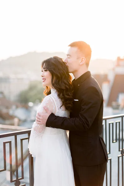 Молода красива Азіатка наречений і наречена на весільній прогулянці, стоячи на терасі будівлі, чоловік обіймаючи жінку зі спини. — стокове фото