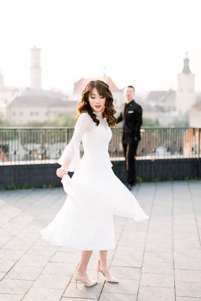 Joven novia asiática hermosa en vestido de novia blanco bailando en la terraza de la antigua ciudad. Novio de pie en el fondo. Paseo de bodas en la ciudad vieja —  Fotos de Stock