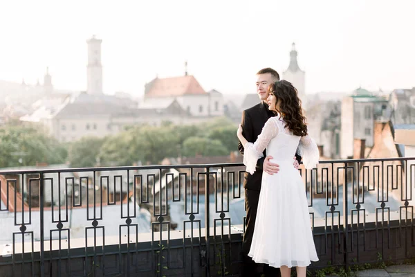 Beautiful Chinese wedding couple embracing and looking at the sunrise while standing against the background of old city panorama